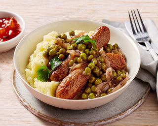 Bowl of food with mashed potatoes, sausages, and peas on a wooden table.