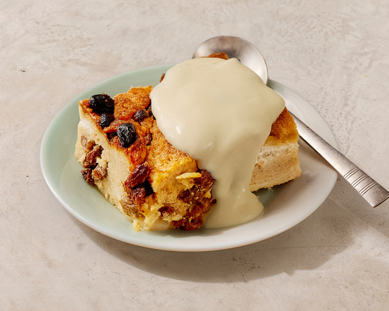 Slice of bread pudding with raisins and a creamy sauce on a plate with a spoon.