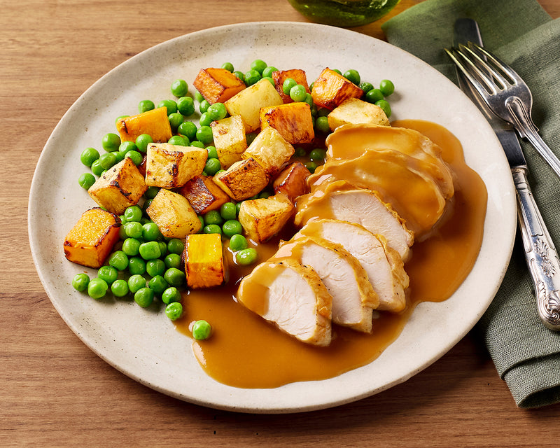 Plated dish with sliced chicken, gravy, roasted potatoes, and peas on a wooden table.