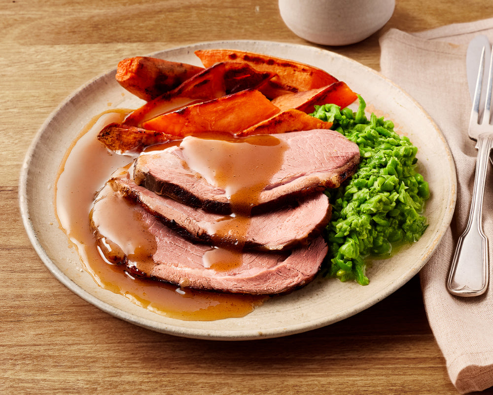 Plated dish of sliced roast lamb with gravy, carrots, and green peas on a wooden table.