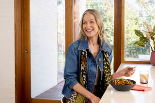 A smiling woman with blonde hair sits at a white table indoors, wearing a blue denim jacket over a patterned top. She is holding a fork next to a bowl of pasta and a glass of water, with large windows and greenery in the background.