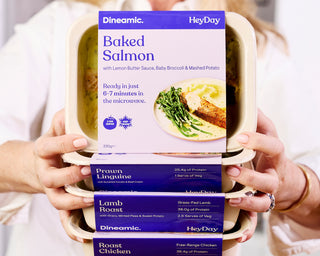 Hands holding a container of Dineamic HeyDay pre-packaged meals with labels on a white background