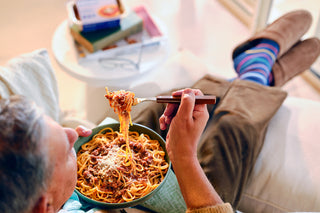 Person eating spaghetti from a bowl with a fork on a couch