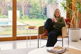 A woman sits in a woven armchair in a bright modern room, holding a mug near large windows that look out onto a garden and pool area. A small side table with books stands beside her, and a potted plant is visible on the right.